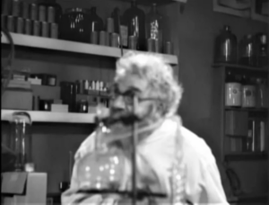 Captura de tela do filme Maniac (1934), em domínio público. A câmera está fora de foco e mostra Horace B. Carpenter como o personagem "Dr. Meirschultz" atrás de um equipamento de laboratório. Captura de tela do filme Maniac (1934), em domínio público. A câmera está fora de foco e mostra Horace B. Carpenter como o personagem "Dr. Meirschultz" atrás de um equipamento de laboratório.