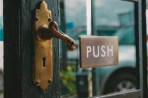 Fotografia de uma porta de madeira pintada de preto com vidro e uma maçaneta alavanca que está sendo virada, de forma a abrir a porta. Ao lado da maçaneta há uma placa onde se lê "Push". Fotografia de uma porta de madeira pintada de preto com vidro e uma maçaneta alavanca que está sendo virada, de forma a abrir a porta. Ao lado da maçaneta há uma placa onde se lê "Push".